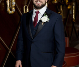 Smiling man in a dark suit with red tie and boutonniere indoors, with saxophones and photos on a patterned wall.