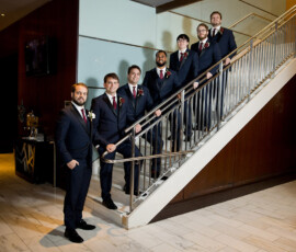 Six men in suits pose on a modern staircase with wood paneling, white tile floors, and contemporary decor.