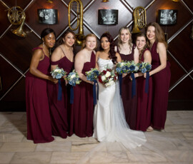 Bride in white with six bridesmaids in burgundy, holding bouquets, indoors by a wall of brass instruments and framed photos.