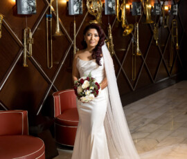 Bride in white gown with bouquet in modern room, gold trombones on wall, framed photos, marble floor, and red chairs.