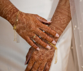 Bride in white dress displays henna-adorned hands with gold jewelry, pink nails, and part of her veil visible.