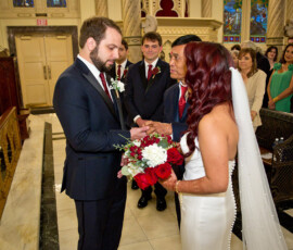 Bride in white with bouquet is given away by her father to the groom in a navy suit, inside a church with watching guests.
