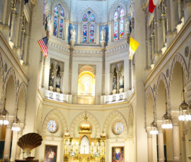 Wedding ceremony in a grand cathedral with stained glass, statues, flags, gold altar, and guests watching the couple.