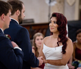 Bride with henna holds groom’s hand during wedding, looking emotional; bridesmaids in burgundy dresses sit behind.