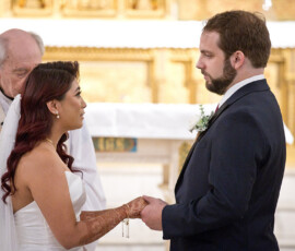 Bride and groom hold hands facing each other at their wedding, officiant in white robes behind them in a decorated church.