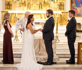Bride and groom hold hands at a church altar with priest, bridesmaid, groomsman, and decorations in the background.