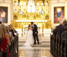 Bride and groom kiss at an ornate church altar, surrounded by guests taking photos. Gold, flowers, and art decorate the scene.