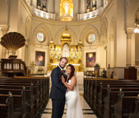 Bride and groom embrace, smiling in an ornate church with stained glass, golden altar, and empty pews; bride holds bouquet.