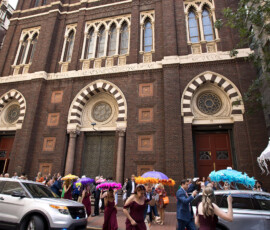 People in burgundy dresses with feathered umbrellas walk by a historic brick building with arched windows and parked cars.