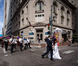 A wedding couple crosses a city street as a band and people with colorful umbrellas parade at a busy intersection.