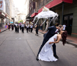 Bride and groom kiss in the street as a parade with a marching band and parasols passes behind them.