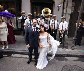 Bride and groom pose on city street with brass band; guests, some with feathered parasols, join the festive scene.