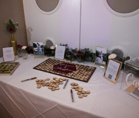 Wedding guest table with pens, sign-in board, wooden hearts, photos, floral décor, and a clear Cards box on a white tablecloth.