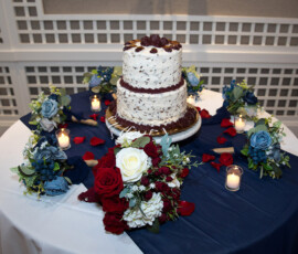 Two-tier cake on a blue and white table, with red, white, and blue flowers, rose petals, and lit candles.