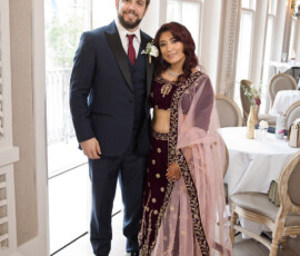 Smiling man in a dark suit and woman in maroon and gold traditional outfit stand together in a bright, elegant room.