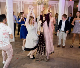 People dance at a formal indoor event; a woman in an ornate traditional dress raises her arms among guests.