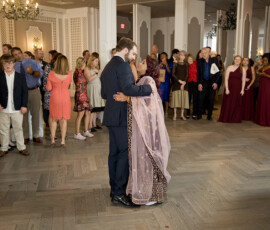 Bride in a pink patterned dress and groom in a dark suit dance at an elegant, festive wedding with smiling guests.