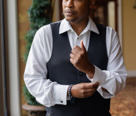 Man in white shirt and black vest adjusts his cuff, gazing thoughtfully out a window indoors on his wedding day.