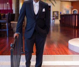 Man in black tuxedo and white shirt stands on indoor stairs with railing; elegant decor, wood floors, and columns behind him.