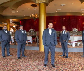 Six men in suits pose for a formal wedding photo in an elegant hall with red curtains and gold columns. Central man stands ahead.
