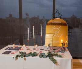 Elegant wedding welcome table with gold mirror sign, candles, invites, rose petals, greenery, and outdoor view.