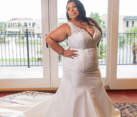 Bride in a white lace wedding dress poses indoors by large windows, with city buildings and palm trees visible outside.