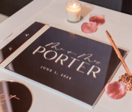 Wedding guest book with “Mr + Mrs Porter, June 1, 2024” in script, rose petals, candles, and pen on white tablecloth.