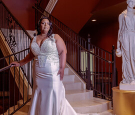 Bride in a white wedding dress on a staircase by a statue, elegant railings, and warm lighting in a formal indoor venue.