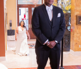 Bride in white gown approaches groom in tuxedo for a romantic indoor first look, with elegant decor and wood flooring.