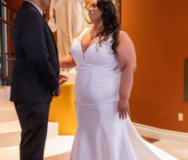 Bride in white gown and groom in dark suit hold hands by staircase, with statue and orange wall background.