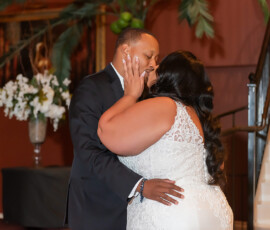 Bride and groom laugh on the dance floor in formal wedding attire—black suit and white lace gown—in an elegant venue.