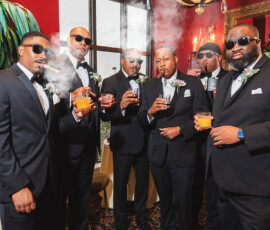 Six men in tuxedos and sunglasses with drinks and cigars pose confidently at a stylish wedding with smoke, red walls, and a chandelier.