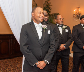 Smiling man in tuxedo with three groomsmen at formal indoor wedding; white drapes and elegant decor in the background.