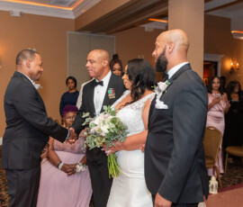 Bride in white gown with two suited men, one shaking hands, at elegant wedding with guests in formalwear watching.