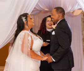 Bride and groom hold hands, smiling at each other under a white wedding arch as the officiant smiles behind them.