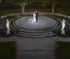 Bride and groom on a circular outdoor platform at night, surrounded by greenery, statues, benches, and soft lighting.