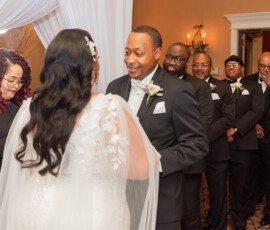 Bride and groom face each other at a warmly lit, elegant wedding with smiling groom, groomsmen in black suits, and officiant nearby.