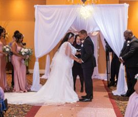 Bride and groom hold hands under a white arch, with pink-clad bridesmaids and groomsmen in black suits at their indoor wedding.