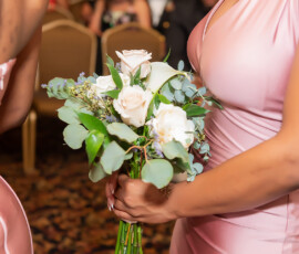 Woman in pink dress holds white and pale pink bouquet; another in similar dress stands nearby at indoor wedding event.
