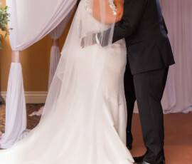Bride and groom in formal attire kiss under white drapery with flower petals on the floor at their wedding ceremony.