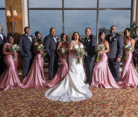 Bride and groom with smiling bridesmaids in pink and groomsmen in dark suits, indoors with large windows behind them.
