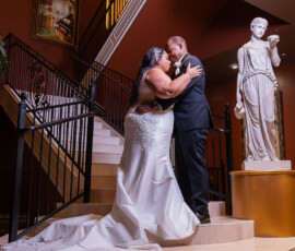 Bride and groom embrace on an elegant staircase beside a tall white statue in a warmly lit indoor wedding setting.
