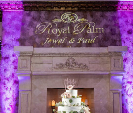 Elegant three-tiered wedding cake with flowers and crown topper, candles, fireplace, and names projected above in script.