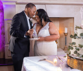 Bride and groom share cake at their wedding reception, with a decorated cake and lit candles on the table, celebrating joyfully.