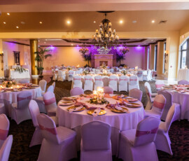 Wedding banquet hall with round tables, white cloths, pink sashes, chandeliers, purple lighting, and floral centerpieces.