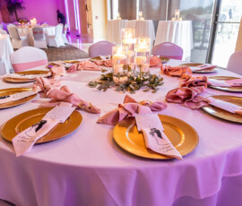 Elegant round table with white cloth, gold chargers, pink napkins, candles, menus, and soft purple-lit wedding décor.