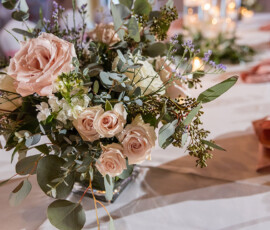 Elegant floral centerpiece with pale pink roses, white blooms, and greenery on a white tablecloth; soft lighting in background.