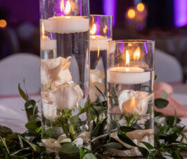 Elegant wedding centerpiece: three glass vases with white roses, floating candles, green leaves, pink napkin, and purple lighting.