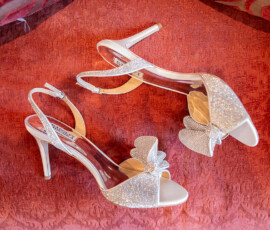 Sparkly open-toe high heels with bows rest on a textured red surface, ideal for weddings or special occasions.