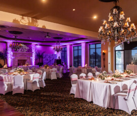 Wedding banquet hall with round tables, white chairs with pink sashes, floral centerpieces, chandeliers, and purple lighting.